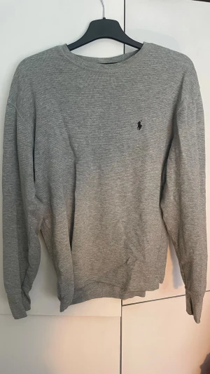 Grå långärmad tröja från Ralph Lauren - Snygg grå långärmad tröja från Ralph Lauren med klassisk rund hals och diskret broderad logga på bröstet. Tröjan är i mjukt, ribbat material och passar perfekt till jeans eller chinos. Det står storlek M men passar även S eller XS ifall man skulle vilja ha den lite oversized. Den är andvänd flera gånger men ser ändå rätt bra ut