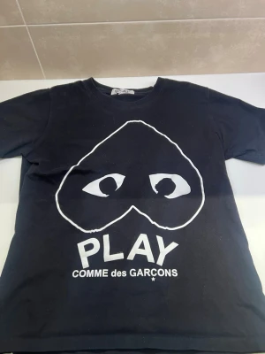 Svart Comme des Garçons Play t-shirt - Cool svart t-shirt från Comme des Garçons Play med det ikoniska hjärtögon-trycket i vitt framtill. Klassisk passform och rund halsringning. Perfekt för dig som gillar streetwear och vill sticka ut med en stilren design.