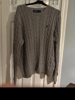 Grå kabelstickad tröja Polo Ralph Lauren - Snygg grå kabelstickad tröja från Polo Ralph Lauren med rund halsringning och broderad logga på bröstet. Tröjan har långa ärmar och ribbade muddar vid ärmslut och nederkant. Perfekt för lager-på-lager och klassisk stil.