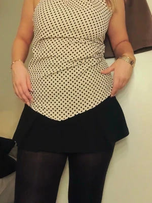 Prickig halternecktop i beige och svart - Snygg halternecktop i beige med svarta prickar. Toppen har en lös passform och är tillverkad i ett lätt, flowy material som känns skönt mot huden. Perfekt att styla med kjol eller jeans för en trendig look.