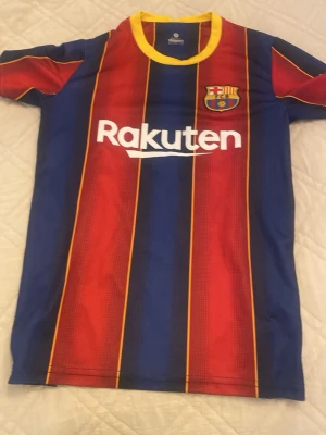 FC Barcelona Ronaldinho matchtröja - Säljer en FC Barcelona matchtröja med Ronaldinho och nummer 10 på ryggen. Tröjan har klassiska blå och röda ränder med gula detaljer, klubbmärke på bröstet och Rakuten som sponsor. Tillverkad i lätt och ventilerande material, perfekt för fotbollsträning eller support.