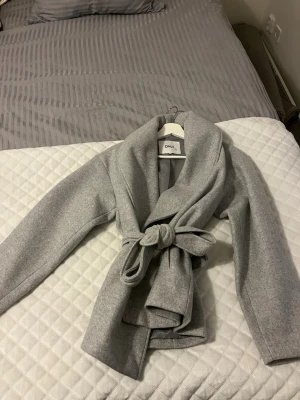 Ljusgrå kappa från ONLY med knytskärp - Stilren ljusgrå kappa från ONLY med bred krage och knytskärp i midjan. Kappan har en mjuk ull-liknande känsla och raka ärmar. Perfekt för dig som gillar minimalistisk och trendig look.
