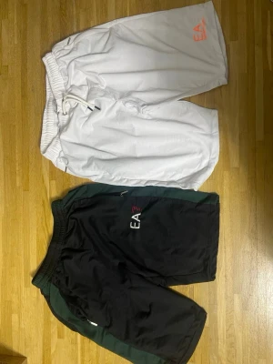 EA7 vita och svarta shorts - Två par shorts från EA7, ett par vita med snörning och ett par svarta med gröna detaljer. Båda har EA7-logga på benet och elastisk midja. Perfekta för en sportig stil och bekväma att bära under varma dagar.