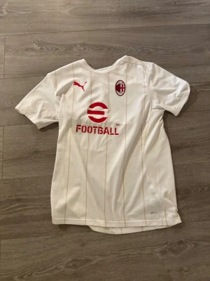 Helt ny omsvänd Vit AC Milan fotbollströja Puma - Helt ny oandvänd Snygg vit AC Milan fotbollströja från Puma med röda detaljer och klubbmärke på bröstet. Tröjan har tunna guldiga ränder, rund hals och är tillverkad i ett lätt och ventilerande material perfekt för träning eller match.
