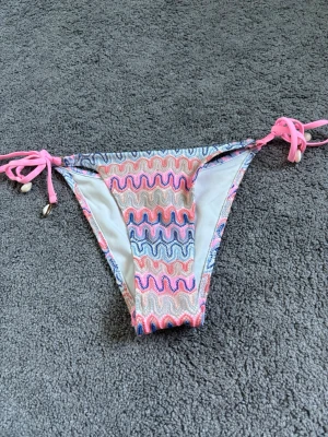 Bikini underdel - Snygga bikinitrosor med vågigt mönster i rosa, blått och vitt. Modellen har knyt i sidorna med rosa snören och små pärldetaljer. Perfekt för strandhäng eller poolparty!