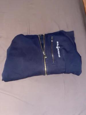 Marinblå hoodie från Sail Racing - Mörkblå hoodie från Sail Racing med dragkedja framtill och en extra bröstficka med dragkedja. Klassisk huva med dragsko och logga på bröstet. Perfekt för dig som gillar sportig och stilren look.