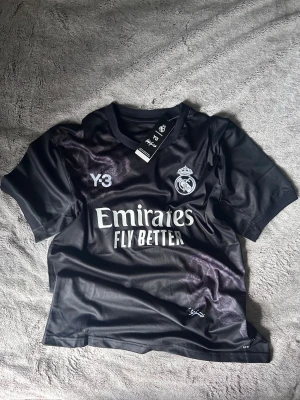 Real Madrid x Y-3 svart matchtröja - Snygg svart Real Madrid matchtröja i samarbete med Y-3. Tröjan har klubbens logga, Y-3 logotyp och 'Emirates Fly Better' tryckt på bröstet. Tillverkad i lätt och ventilerande material, perfekt för fotboll eller träning. Rund halsringning och korta ärmar.