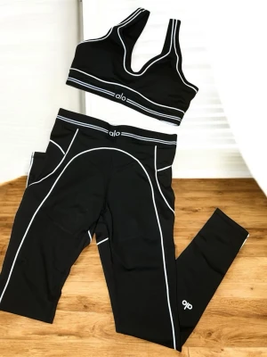 Svart träningsset från Alo Yoga - Snyggt svart träningsset från Alo Yoga med sport-bh och tights. Setet har vita kontrastsömmar och logga på både bh och byxa. Tightsen har hög midja och praktiska fickor på sidorna. Perfekt för gymmet eller yoga.