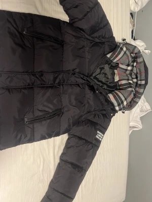 Svart pufferjacka från Burberry - Snygg svart pufferjacka från Burberry London med klassiskt rutigt foder i grått, vitt och rött. Jackan har dragkedja, två snedställda fickor med dragkedja och en huva med rutigt innerfoder. Perfekt för kalla vinterdagar och riktigt trendig.
