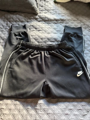 Svarta Nike byxor  - Svarta mjukisbyxor från Nike med vit logga på vänster ben och vita detaljer längs sidorna.. Perfekta för en avslappnad stil och riktigt sköna att ha på sig.