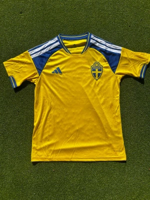 Svensk landslagströja Adidas gul/blå - Gul och blå fotbollströja från Adidas med Sveriges landslagsemblem på bröstet. Tröjan har korta ärmar, rund hals och klassiska Adidas-ränder på axlarna. Perfekt för match eller träning. Materialet är lätt och andas. 68cm längd och 48cm bredd