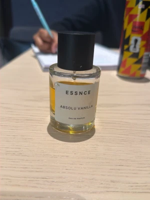 ESSNCE Absolu Vanilla parfym - En stilren parfymflaska från ESSNCE med doften Absolu Vanilla. Flaskan är genomskinlig med svart lock och innehåller en gyllengul vätska. Minimalistisk etikett framtill. Perfekt för dig som gillar varma, söta doftnoter.