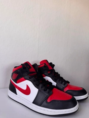 Nike Air Jordan 1 Mid röd/svart/vit - Snygga Nike Air Jordan 1 Mid sneakers i rött, svart och vitt läder. Klassisk siluett med svart snörning, röd swoosh och Air Jordan-logga på sidan. Perfekta för dig som gillar streetwear och vill sticka ut med en ikonisk modell.