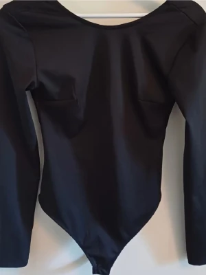 Svart långärmad body - Snygg svart body med långa ärmar och djup rygg. Tillverkad i ett stretchigt, slätt material som sitter tight mot kroppen. Perfekt att styla med jeans eller kjol för en clean look. 