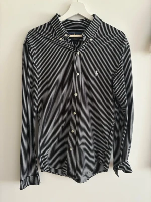 Randig skjorta från Ralph Lauren - Snygg svart skjorta med vita ränder från Ralph Lauren. Slim fit-modell med klassisk krage och knäppning framtill. Ikoniska loggan broderad på bröstet. Perfekt för dig som gillar stilrena och tidlösa plagg. Endast använd fåtal gånger!