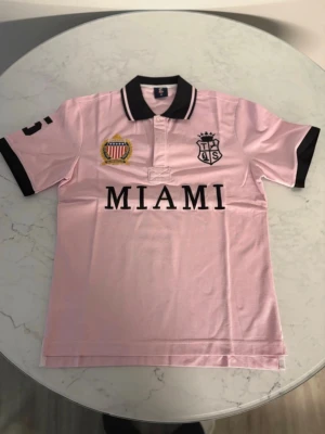 Ljusrosa piké med MIAMI-tryck - Snygg ljusrosa pikétröja med svart krage och ärmslut. Framtill står det MIAMI i stora bokstäver och två broderade märken pryder bröstet. Tröjan har korta ärmar och klassisk pikéknäppning. Perfekt för en sportig och trendig look.