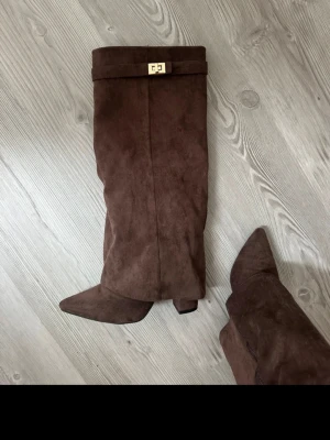 Bruna knähöga stövlar i mocka-look 38 - Snygga bruna knähöga stövlar med spetsig tå och klack. Tillverkade i mockaimitation med dragkedja på insidan och dekorativt spänne upptill i guld. Perfekta för att lyfta din outfit med en trendig touch.