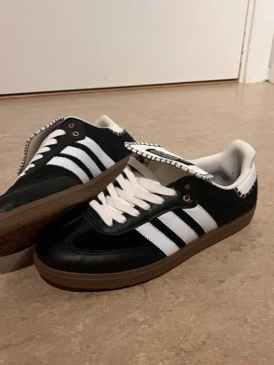 Adidas Wales Bonner svartvita sneakers - Snygga Adidas Wales Bonner sneakers i svart och vitt med klassiska tre ränder på sidan och unik detalj på plösen. Skorna har vit insida, vita skosnören och brun gummisula. Materialet är en mix av läder och textil, perfekt för dig som gillar streetstyle.