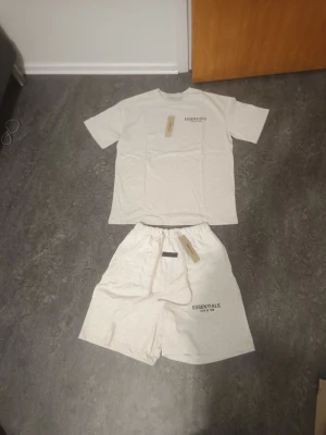 Essentials Fear of God set  - Säljer ett snyggt tvådelat set Essentials Fear of God i beige. Setet består av en t-shirt med rund hals och tryck fram och bak samt matchande shorts med resår i midjan och tryck på benet. Båda delar i mjukt bomullsmaterial, perfekt för chill dagar.