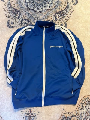 Blå Palm Angels ziptröja - Snygg blå ziptröja från Palm Angels med vita ränder längs ärmarna och vit dragkedja. Tröjan har hög krage, fickor med dragkedja och logga på bröstet. Perfekt för en sportig och trendig look.