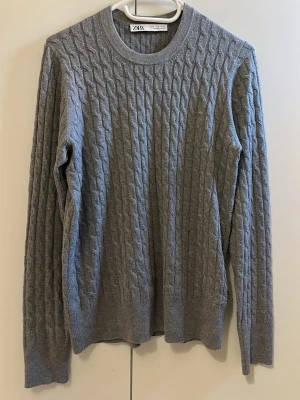 Grå kabelstickad tröja från Zara - Snygg grå kabelstickad tröja från Zara med rund halsringning och långa ärmar. Tröjan har ett klassiskt flätmönster och är perfekt för lager-på-lager-stil. Passar dig som gillar enkel och stilren design.