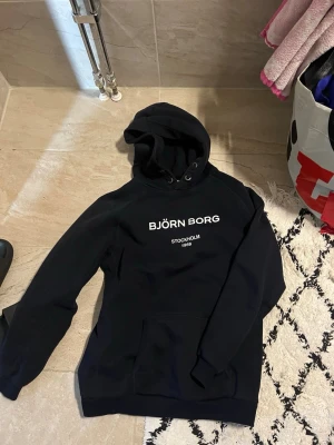 Svart hoodie från Björn Borg - Svart hoodie från Björn Borg med vit text på bröstet där det står 'BJÖRN BORG STOCKHOLM 1984'. Klassisk modell med huva och känguruficka framtill. Perfekt för en avslappnad och sportig stil.