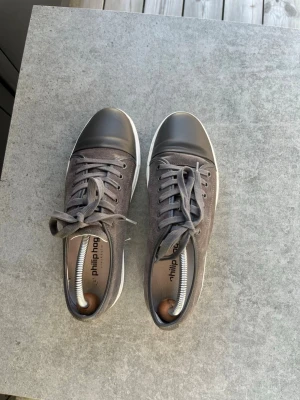 Grå sneakers från Philip Hog - Snygga grå sneakers från Philip Hog med mockaliknande ovandel och mörkgrå tåparti i läder. Skorna har vita sulor och matchande grå skosnören. Perfekta för en stilren och avslappnad look. 