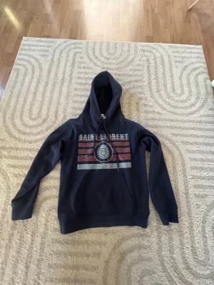 Mörkblå Saint Laurent hoodie - Snygg mörkblå hoodie från Saint Laurent med tryck framtill i vitt, rött och grått. Klassisk känguruficka och justerbar huva med snören. Perfekt för dig som gillar streetwear och vill ha en hoodie med coolt tryck och skön passform.