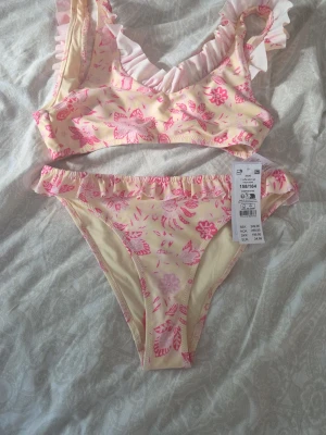 Blommig bikini Från gina 158/164 - Söt bikini från Gina tricot i ljusgul med rosa blommönster och volangdetaljer vid axelband och midja. perfekt för strand eller pool. Materialet är stretchigt och snabbtorkande. Aldrig använd eller testad då det blev fel bikini💕 