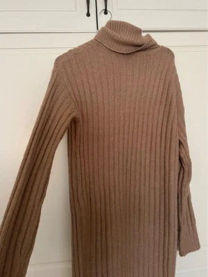 Beige ribbad stickad klänning - Mysig beige stickad klänning med polokrage och lång ärm. Klänningen är ribbad och har en rak passform som ger en avslappnad look. Perfekt för kyligare dagar och enkel att styla med boots eller sneakers.