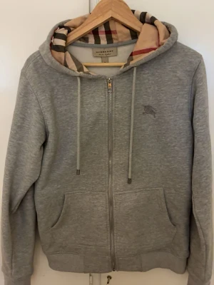 Grå zip-hoodie från Burberry - Snygg grå hoodie med dragkedja från Burberry. Klassisk huva med det ikoniska rutmönstret på insidan, dragsnören och två stora fickor framtill. Broderad logga på bröstet. Perfekt för en chill och stilren look.