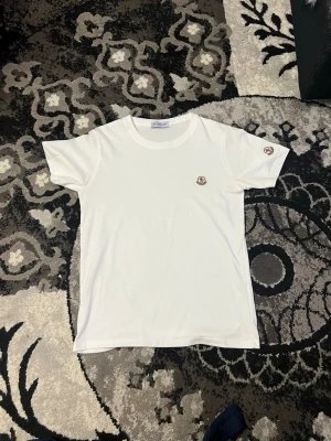 Vit Moncler t-shirt med logga - Snygg vit t-shirt från Moncler med klassisk rund halsringning och liten broderad logga på bröstet samt ärmen. Perfekt basic-plagg med stilren design som passar till det mesta. Tillverkad i mjuk bomull för skön känsla.