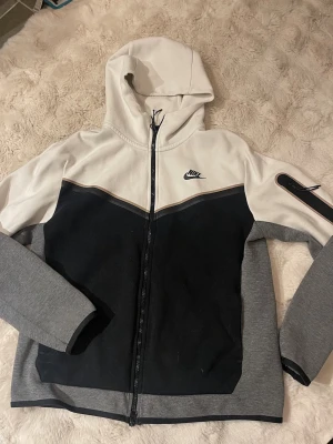 Nike hoodie i svart, grå och vit - Snygg hoodie från Nike med dragkedja, huva och färgblock i svart, grått och vitt. Klassisk Nike-logga på bröstet och en cool detalj på ena ärmen. Perfekt för en sportig och avslappnad stil.