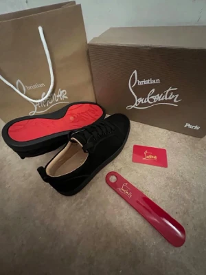 Christian Louboutin svarta sneakers - Snygga svarta sneakers från Christian Louboutin med klassisk röd sula och diskret logga. Skorna har en låg profil, rund tå och är tillverkade i mocka med svarta skosnören. Perfekt för dig som vill ha en exklusiv och stilren look. Kommer också med kvitto 