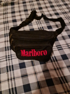 Svart Marlboro magväska - Svart magväska i tyg med röd Marlboro-logga framtill. Väskan har flera dragkedjefack och justerbart midjeband med spänne. Perfekt för att bära småsaker och hålla händerna fria. Klassisk och stilren design.