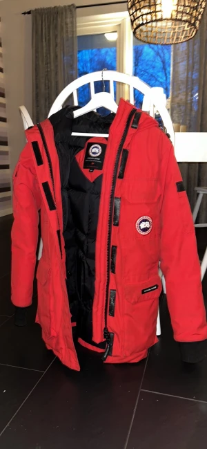 Röd parkas från Canada Goose - Röd parkasjacka från Canada Goose med svart foder och klassisk logga på bröstet. Jackan har huva med fuskpäls, flera fickor och svarta detaljer längs dragkedjan. Perfekt för kalla dagar och riktigt snygg streetstil.