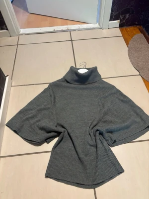 Stickad poncho med polokrage - Mysig grå stickad poncho med breda ärmar och hög polokrage. Perfekt att slänga över en långärmad tröja för extra värme. Enkel och stilren design som passar till det mesta. Materialet känns mjukt och bekvämt. Aldrig använd! 