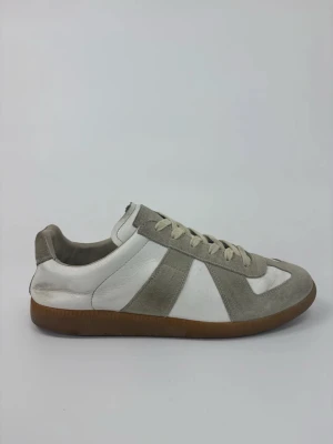 Vita och grå Maison Margiela sneakers med gummisula - Snygga sneakers i vitt och ljusgrått med klassisk design. Ovandelen är i en mix av läder och mocka, och sulan är i brunt gummi för en retrovibe. Perfekta för dig som gillar stilrena och tidlösa skor. Skorna har lågt skaft och neutrala färger som passar till det mesta.