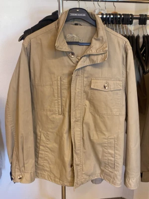 Beige jacka från Rappson  - Stilren beige jacka från Rappson med klassisk krage och flera fickor framtill, inklusive en bröstficka med knapp. Jackan har dragkedja och knappar framtill samt långa ärmar. Perfekt för dig som gillar enkel och tidlös stil.