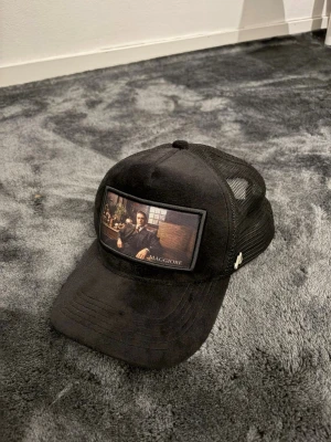 Svart keps från Maggiore med motiv - Snygg svart keps från Maggiore med mesh på sidorna och ett motiv framtill som föreställer en klassisk filmscen. Kepsen har böjd skärm och justerbar baksida. Perfekt accessoar för en cool streetstyle-look.