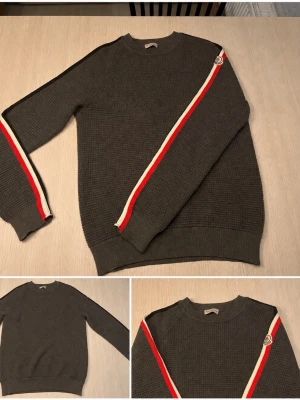 Mörkgrå stickad tröja Moncler - ÄKTA✅ Snygg och sällsynt mörkgrå stickad tröja från Moncler med vita och röda ränder längs ärmarna. Tröjan har rund halsringning, ribbade muddar och ett Moncler-märke på ena ärmen. Perfekt för dig som gillar stilren design.