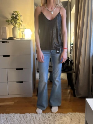 Svart glittrig linnetopp med smala band - Snygg svart glittrig linnetopp med tunna axelband och djup v-ringning. Toppen har en lös passform och är perfekt för att skapa en trendig look. Materialet är glansigt och ger en cool effekt.