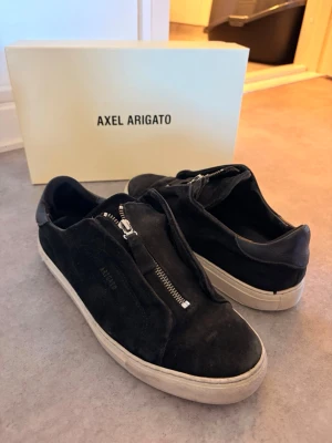 Svarta Axel Arigato sneakers med dragkedja - Snygga svarta sneakers från Axel Arigato i mocka med unika dragkedjor istället för klassiska snören. Skorna har vit sula och låg profil, perfekt för en stilren och modern look. Märkeslogga diskret på sidan. Kommer med originalkartong.