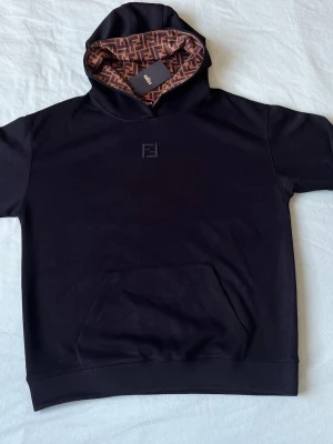 Svart hoodie från Fendi med FF-mönster - Svart hoodie från Fendi med korta ärmar och klassisk känguruficka. Insidan av huvan har det ikoniska bruna FF-mönstret. Liten Fendi-logga broderad på bröstet. Perfekt för dig som gillar lyxiga streetwear-plagg och vill sticka ut.