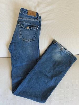 Blå raka jeans med fickdetaljer - Säljer ett par klassiska blå jeans med raka ben och snygga fickdetaljer bak. Jeansen har en ljus tvätt och en liten läderpatch i midjan. Perfekta till vardagslooken och passar till det mesta. Materialet är denim i bomull.