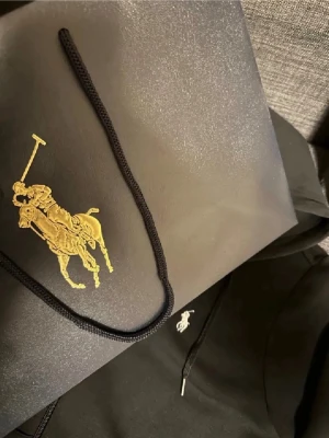 Svart hoodie från Polo Ralph Lauren - Snygg svart hoodie från Polo Ralph Lauren med dragkedja, huva och klassiska vita loggan broderad på bröstet. Hoodien har fickor framtill och justerbara snören i huvan. Perfekt för en chill och stilren look.