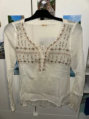 Vit boho-blus med broderi och pärlor från Hollister - Supersöt vit blus i tunn bomull med långa ärmar, broderade detaljer och små pärlor framtill. Djup v-ringning med snörning ger en bohemisk vibe. Perfekt till jeans eller kjol för en chill look.