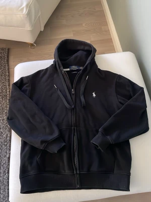 Svart hoodie från Polo Ralph Lauren - Svart zip tröja med dragkedja från Polo Ralph Lauren i storlek S. Klassisk modell med huva, dragsnören och vit broderad logga på bröstet. Custom fit-passform och ribbade muddar vid ärmslut och nederkant. Perfekt för en avslappnad och stilren look. Inga fläckar eller liknande, den är använd 1-2 gånger men passar inte längre.