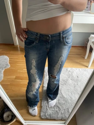 Super low waist bootcut jeans  - Säljer ett par blå jeans med slitna detaljer och hål på båda benen. Jeansen har låg midja, klassisk femficksmodell Perfekta för en avslappnad och trendig look. 