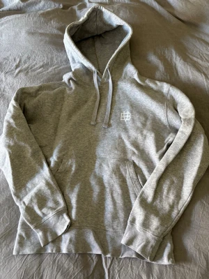 Grå hoodie från Bvaldi med logga - Snygg grå hoodie från Bvaldi med vit broderad logga på bröstet. Klassisk modell med huva, dragsnören och magficka. Mjuk insida och ribbade muddar vid ärmslut och nederkant. Perfekt för en chill och avslappnad stil.